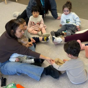 An adult and several young children sit in a circle on the floor, interacting and sharing items from a brown paper bag during a group activity.