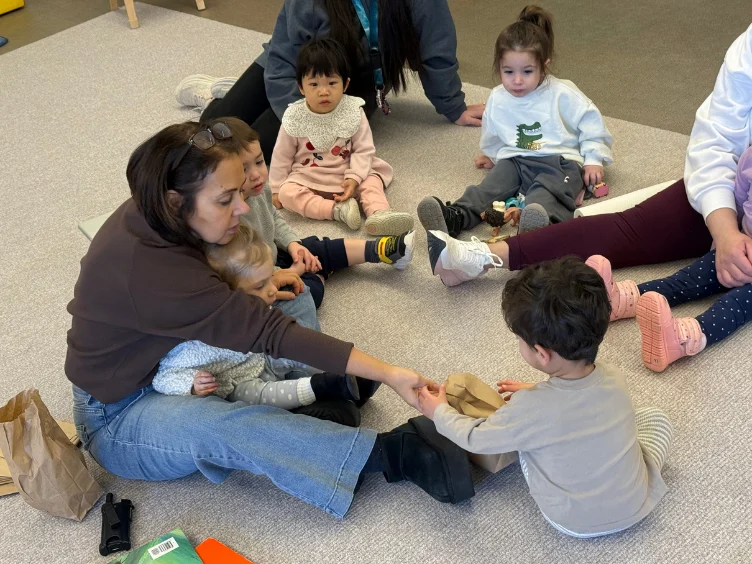 An adult and several young children sit in a circle on the floor, interacting and sharing items from a brown paper bag during a group activity.