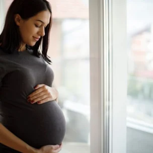 Pregnant woman standing indoors by a window, gently holding her belly and looking down with a slight smile.