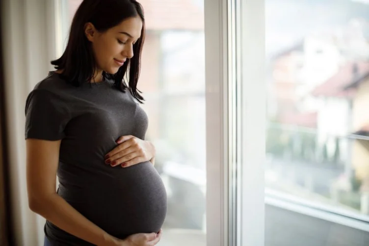Pregnant woman standing indoors by a window, gently holding her belly and looking down with a slight smile.