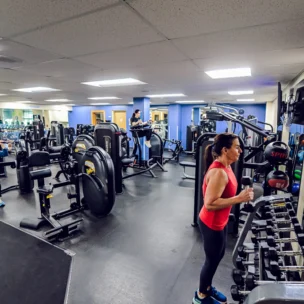 People working out in a gym with various exercise machines and equipment; one person lifts dumbbells by a mirror, others use cardio and strength machines.