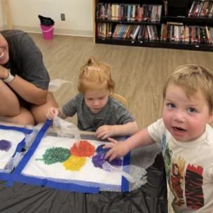 Two young children use bubble wrap for a colorful painting activity at a table while an adult woman smiles and watches. Bookshelves and a pink bucket are visible in the background.