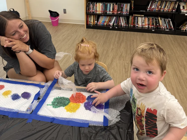Two young children use bubble wrap for a colorful painting activity at a table while an adult woman smiles and watches. Bookshelves and a pink bucket are visible in the background.
