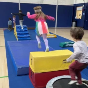 Children participate in an indoor obstacle course, jumping on mats and trampolines in a gymnasium with blue walls and a wooden floor.