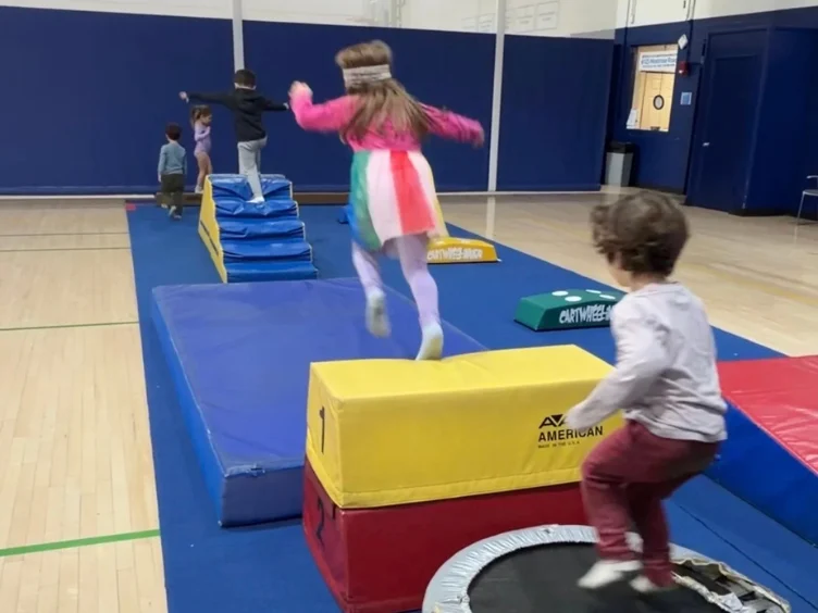 Children participate in an indoor obstacle course, jumping on mats and trampolines in a gymnasium with blue walls and a wooden floor.
