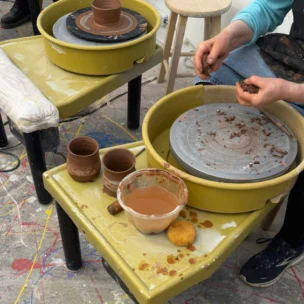 A person shapes clay at a pottery wheel with finished clay cups, a bowl of water, tools, and a sponge on a messy, paint-splattered floor.