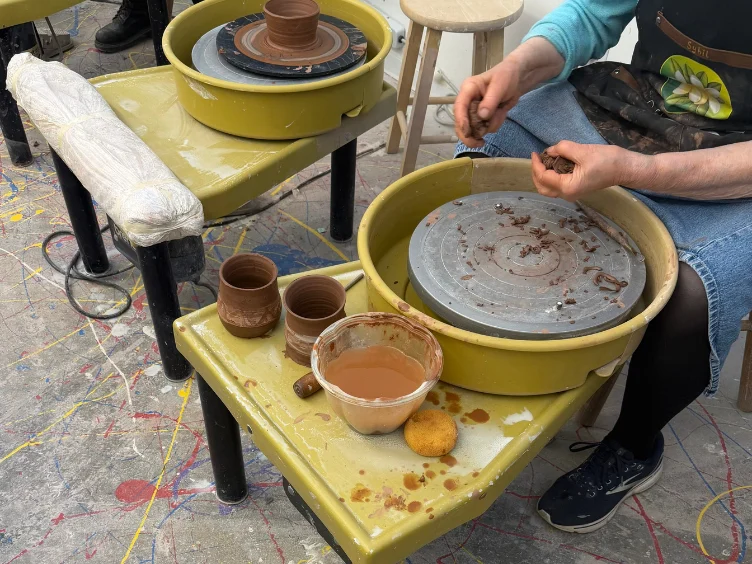 A person shapes clay at a pottery wheel with finished clay cups, a bowl of water, tools, and a sponge on a messy, paint-splattered floor.