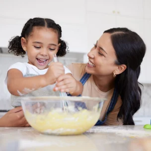 An adult and a young girl smile while mixing batter in a glass bowl in a bright kitchen, surrounded by baking tools.