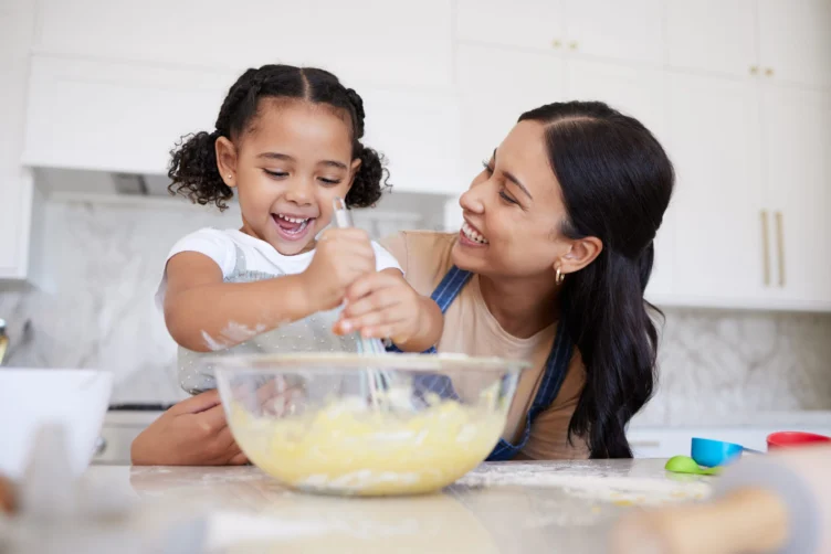 An adult and a young girl smile while mixing batter in a glass bowl in a bright kitchen, surrounded by baking tools.