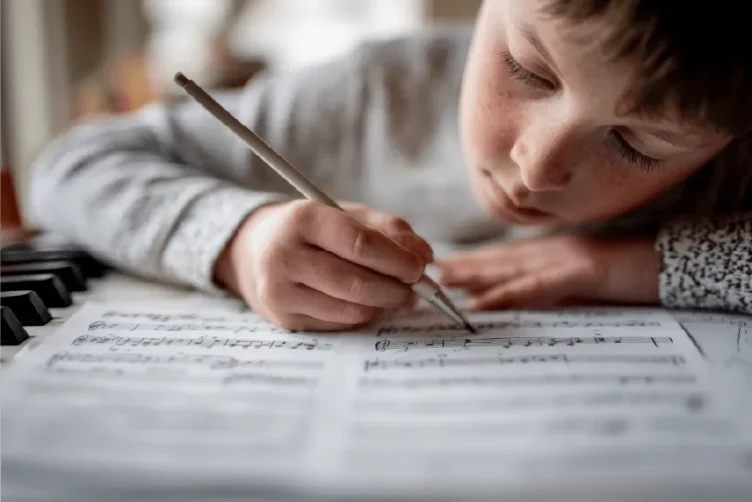 A child writes with a pencil on a sheet of music, with part of a keyboard visible beside the paper.