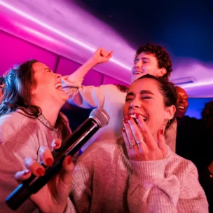 Five people sing and laugh together in a karaoke room with colorful lighting. One woman holds a microphone, and the group appears joyful and energetic.