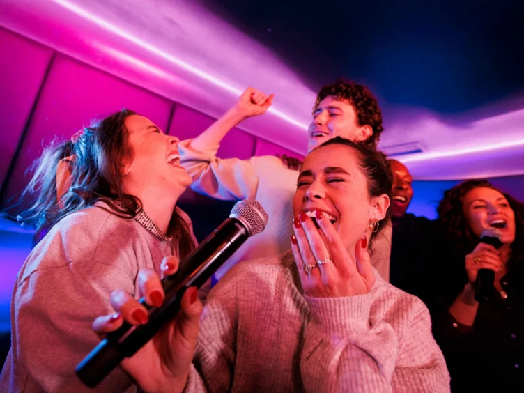 Five people sing and laugh together in a karaoke room with colorful lighting. One woman holds a microphone, and the group appears joyful and energetic.