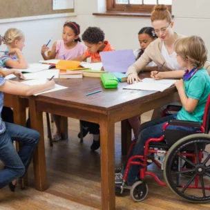 A teacher sits at a table with a diverse group of children, including one in a wheelchair, all working on school assignments in a classroom.