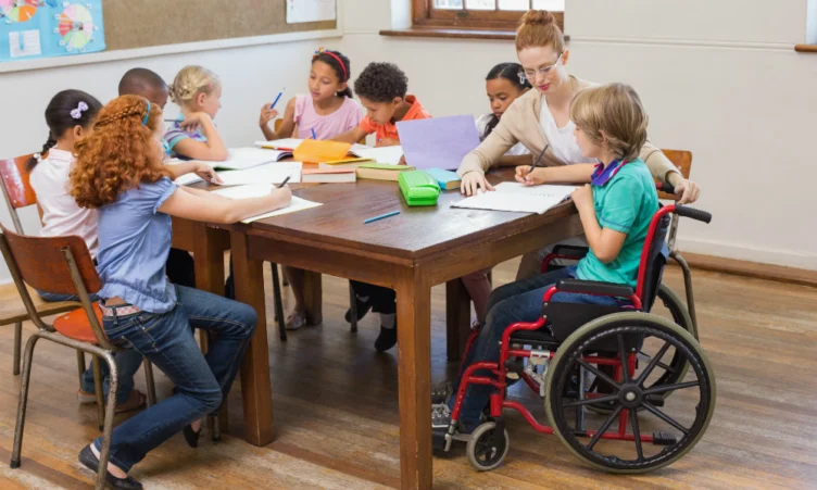 A teacher sits at a table with a diverse group of children, including one in a wheelchair, all working on school assignments in a classroom.