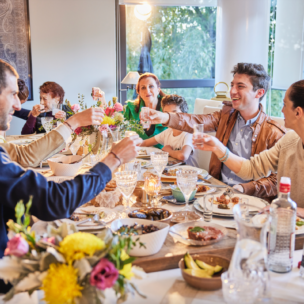 A group of people sit around a dining table sharing a meal and toasting drinks, with food, flowers, and drinks on the table in a well-lit room.
