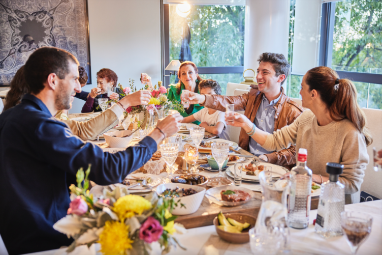 A group of people sit around a dining table sharing a meal and toasting drinks, with food, flowers, and drinks on the table in a well-lit room.