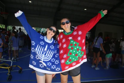 Two women in sunglasses wear a split holiday sweater—one side featuring a blue Hanukkah menorah and stars, the other side featuring a red Christmas tree—standing in a gymnasium.