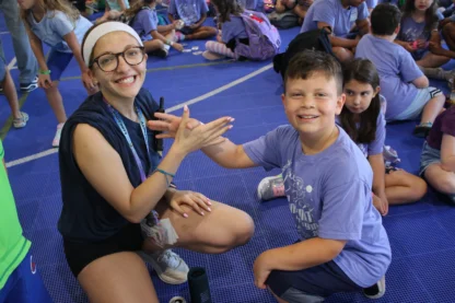 A smiling adult and a smiling boy in matching purple shirts sit on a gym floor, clasping hands, with other children seated in the background.