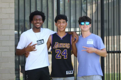 Three young men stand together smiling; one wears a Lakers jersey, the others wear casual shirts. They gesture with peace signs in front of a metal fence and brick wall.