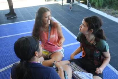 Three young people sit in a circle on a blue mat, talking and smiling, with a wheelchair and greenery visible in the background.