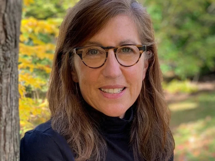 Woman with long brown hair and glasses wearing a black turtleneck, standing outdoors near a tree, with blurred greenery in the background.