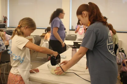 Two girls mix ingredients with a hand mixer in a large bowl at a table while another person stands nearby in a classroom setting, enjoying hands-on activities commonly found at a summer camp near me.