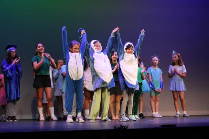 Children in costumes, including three in shark suits, stand on stage with others in various outfits, clapping and smiling under stage lights—capturing the joyful spirit you’ll find at a summer camp near me.