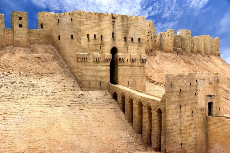 Ancient stone fortress with arched entrance and high walls, built on a sandy hill under a partly cloudy blue sky.