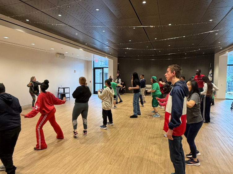 A group of people in casual clothes participate in a dance or exercise class in a spacious indoor room with wood flooring and a high ceiling.
