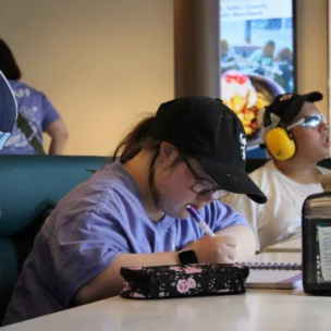 A person wearing a black cap and glasses writes in a notebook at a table in a casual indoor setting, with others seated nearby.
