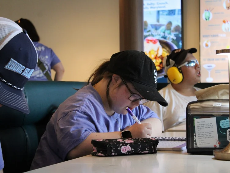 A person wearing a black cap and glasses writes in a notebook at a table in a casual indoor setting, with others seated nearby.