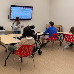 Seven people sit at tables in a classroom, facing a screen displaying a presentation. One person stands in front, presenting to the group.