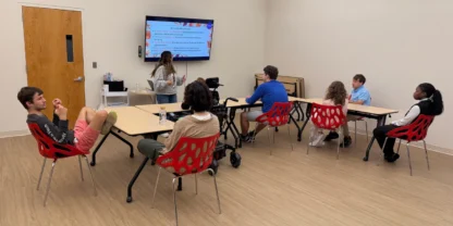 Seven people sit at tables in a classroom, facing a screen displaying a presentation. One person stands in front, presenting to the group.