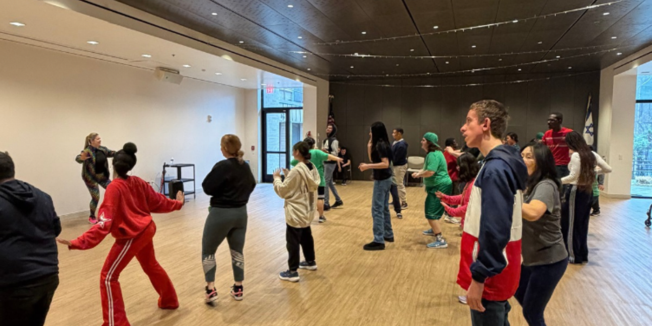 A group of people participates in a dance or fitness class in a spacious room with wooden floors and large windows.