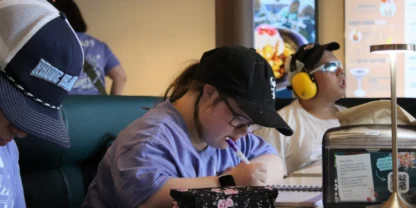 Three people sit at a table; one writes in a notebook, another wears headphones and looks up, and a third person sits nearby. A menu board is visible in the background, adding to the cozy atmosphere reminiscent of planning sessions at a summer camp near me.