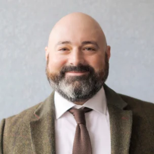 A bald man with a beard, wearing a white shirt, brown tie, and a patterned blazer.