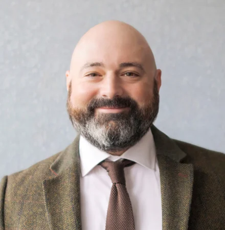 A bald man with a beard, wearing a white shirt, brown tie, and a patterned blazer.