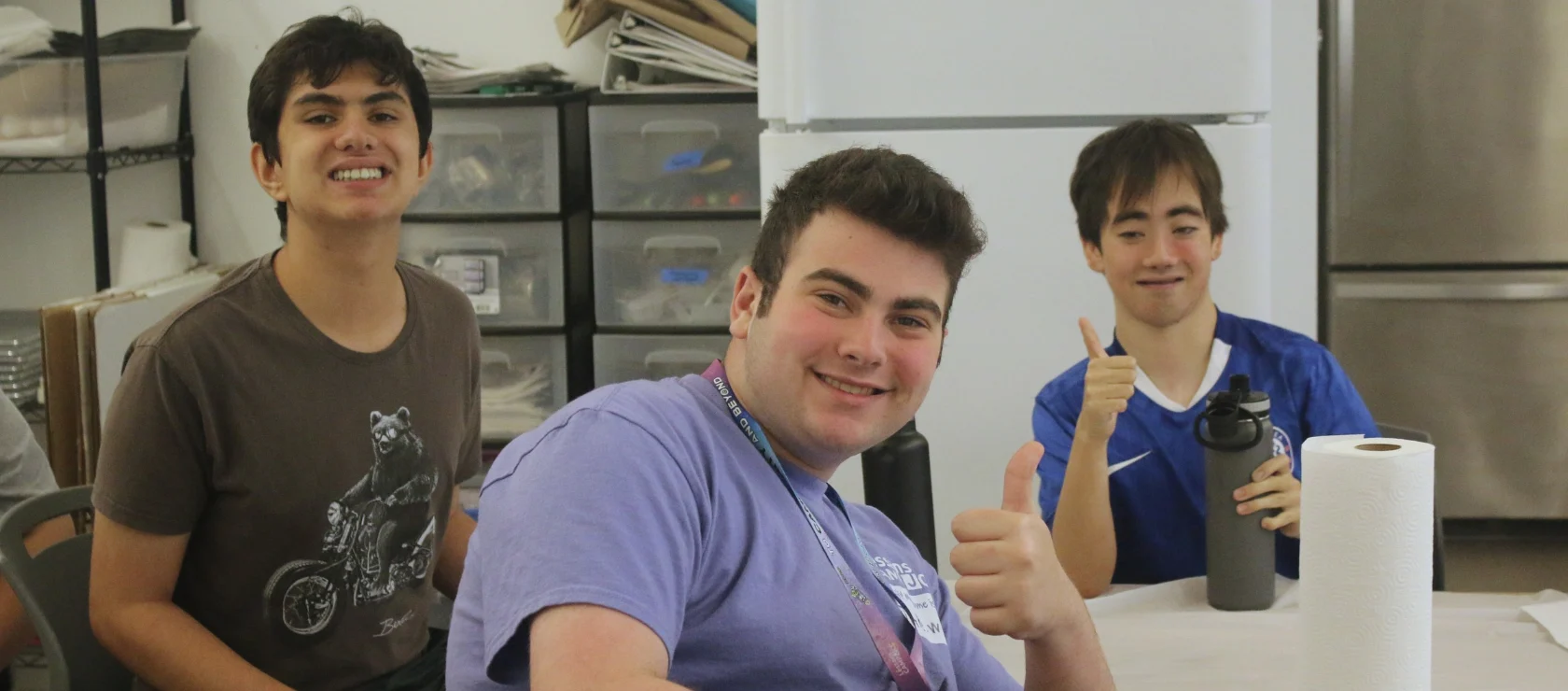 Three young men smiling at a table, two giving a thumbs-up.
