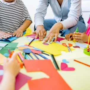 An adult and three children sit around a table working on a paper craft project with scissors, glue, and colored construction paper.
