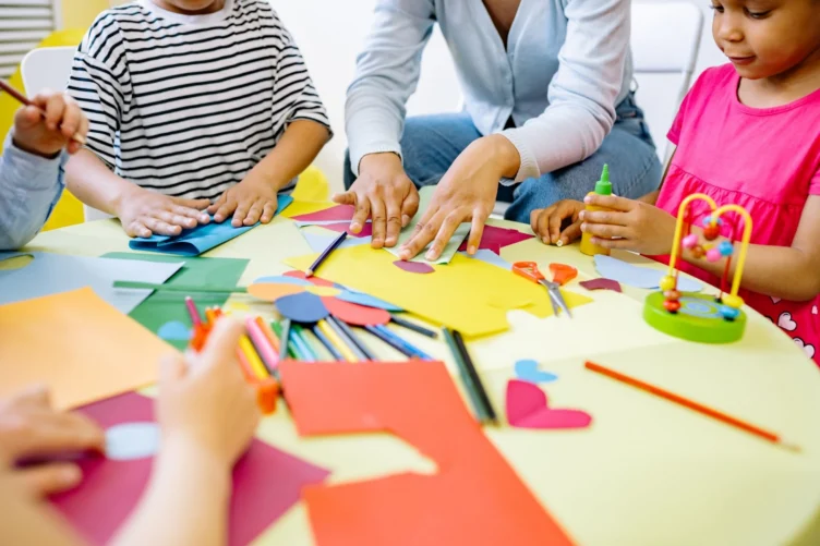 An adult and three children sit around a table working on a paper craft project with scissors, glue, and colored construction paper.