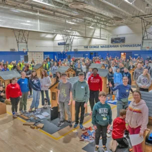 A large group of volunteers posing in a gymnasium around several small, unfinished wooden dog houses.