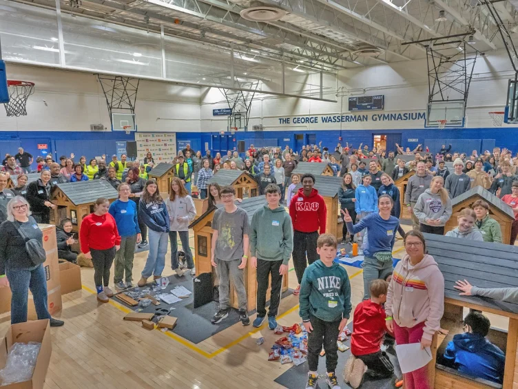 A large group of volunteers posing in a gymnasium around several small, unfinished wooden dog houses.