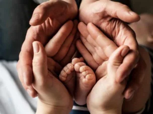 Four adult hands cupped together protectively around a newborn baby’s feet.