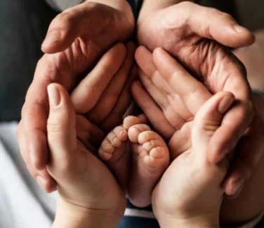Four adult hands cupped together protectively around a newborn baby’s feet.