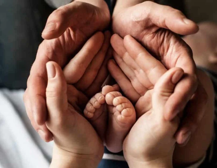 Four adult hands cupped together protectively around a newborn baby’s feet.