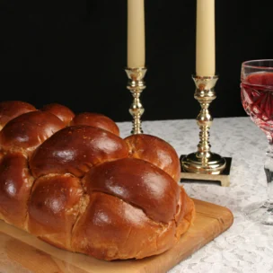 A loaf of braided challah bread on a wooden board next to two lit candles and a glass of red wine.
