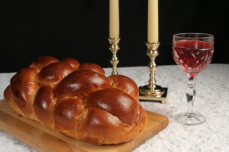 A loaf of braided challah bread on a wooden board next to two lit candles and a glass of red wine.