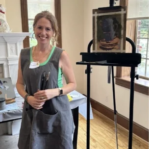 A smiling woman wearing a grey apron and holding paintbrushes stands next to an easel displaying a still-life painting of cups.