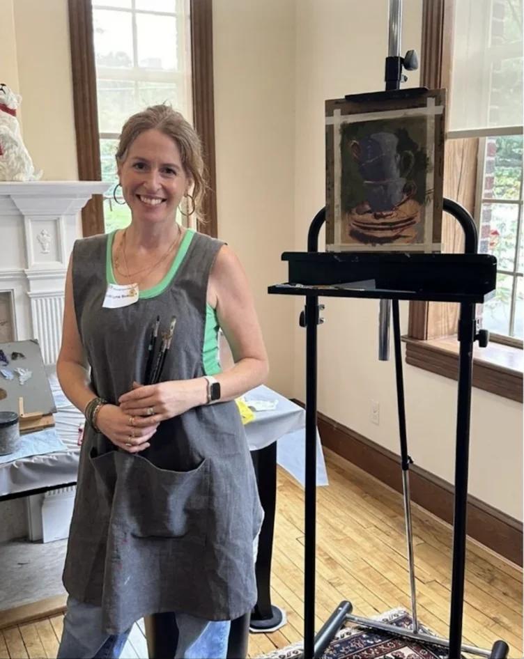 A smiling woman wearing a grey apron and holding paintbrushes stands next to an easel displaying a still-life painting of cups.