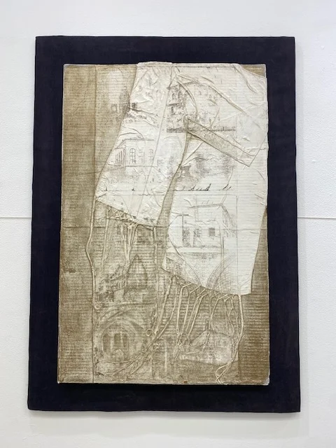 Mixed-media collage featuring layered paper with architectural sketches and hanging fabric threads on a dark rectangular backing.